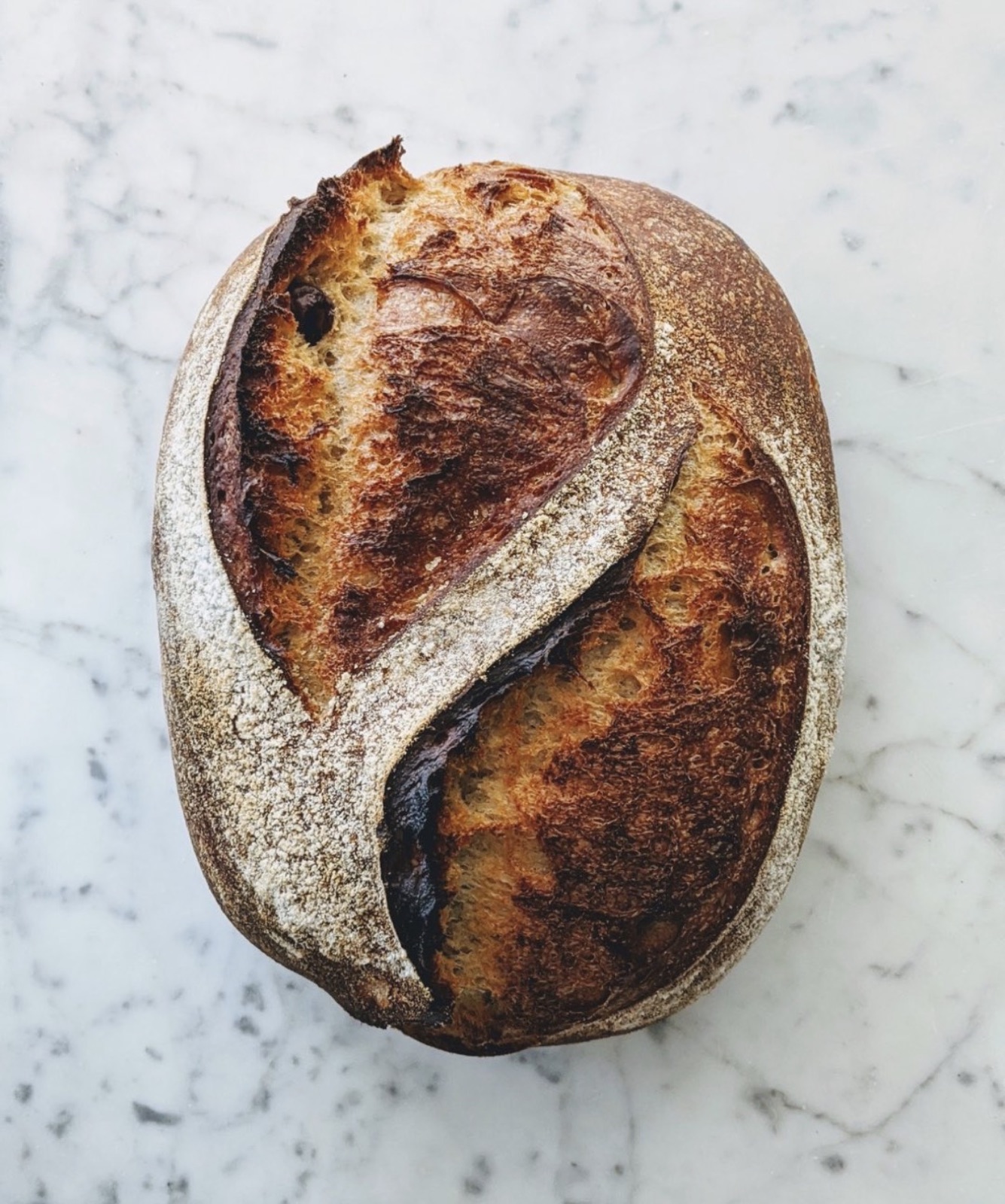 A dark-crusted sourdough boule on marble, scored in two crescent slashes.