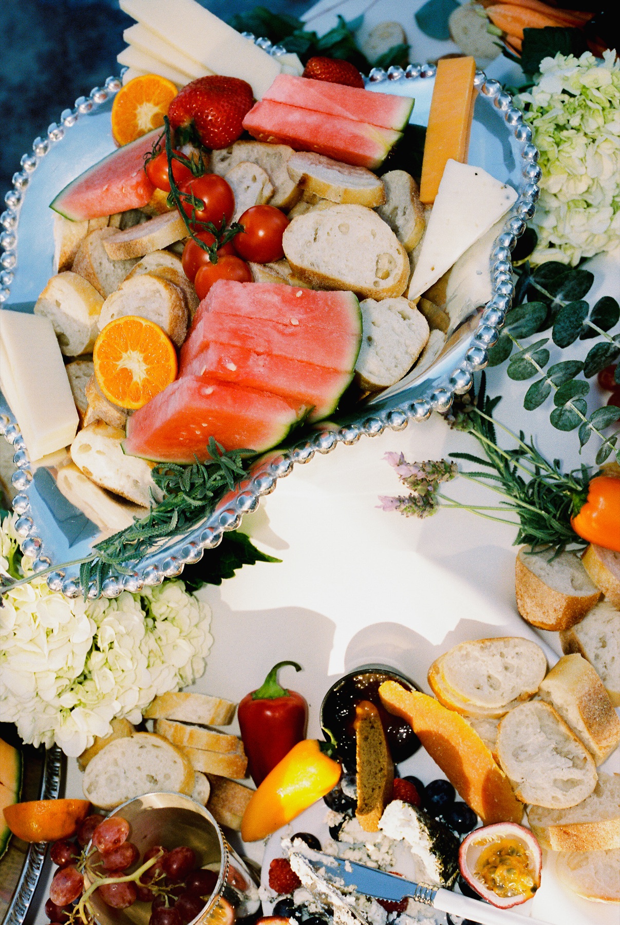 A Mangia Farms grazing tablescape on beaded silver platters: sliced sourdough and baguette, watermelon, citrus, cheeses, cherry tomatoes, grapes, herbs and florals.